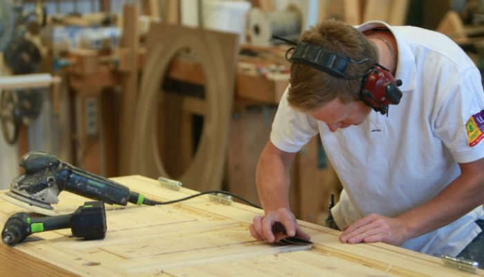 A male carpenter in white t-shirt sanding a wooden door.